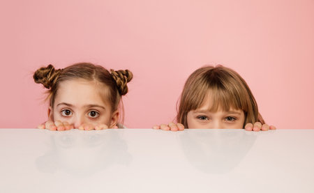 Happy kids, girls on coral pink studio background. Look happy, cheerful. Copyspace for ad. Childhood, education, emotions, business, facial expression concept. Peeking out from behind the tableの写真素材