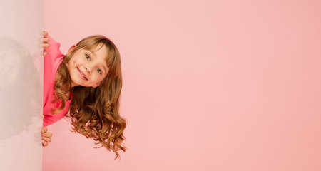 Happy kid, girl isolated on coral pink studio background. Looks happy, cheerful. Copyspace for ad. Childhood, education, emotions, facial expression concept. Peeking out from behind the wall. Flyerの写真素材