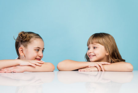 Happy kids, girls isolated on blue studio background. Look happy, cheerful. Copyspace for ad. Childhood, education, emotions, business, facial expression concept. Sitting at the table, laughtingの写真素材