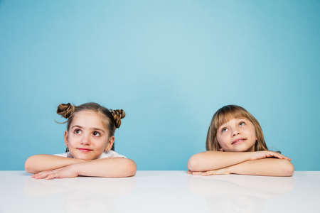 Happy kids, girls isolated on blue studio background. Look happy, cheerful. Copyspace for ad. Childhood, education, emotions, business, facial expression concept. Sitting at the table, dreamingの写真素材