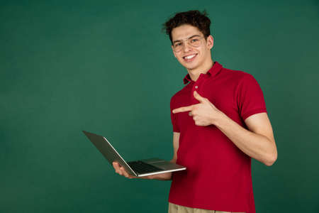 Pointing on screen. Caucasian handsome mans portrait isolated on green studio background with copyspace. Male model with laptop. Concept of human emotions, facial expression, sales, ad, fashion.の写真素材