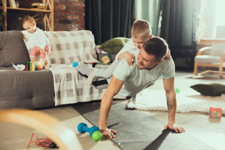 Plank. Young man exercising fitness, aerobic, yoga at home, sporty lifestyle. Getting active while his child playing on the background, home gym. Healthcare, movement, parenthood concept.の写真素材