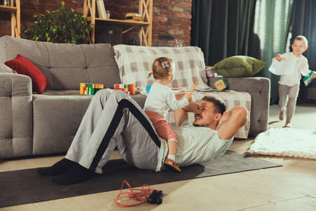 ABS workout. Young man exercising fitness, aerobic, yoga at home, sporty lifestyle. Getting active while his child playing on the background, home gym. Healthcare, movement, parenthood concept.の写真素材