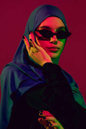 Style. Beautiful arab woman in stylish hijab isolated on burgundy studio background in neon light...