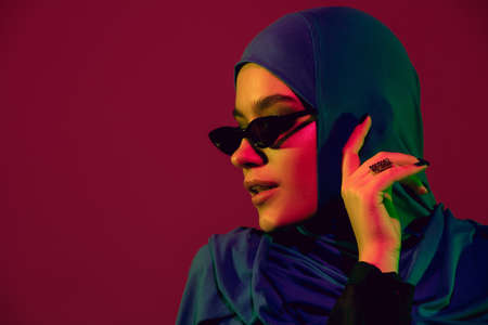 Style. Beautiful arab woman in stylish hijab isolated on burgundy studio background in neon light with copyspace for ad. Fashion, beauty, style concept. Female model with trendy make up, accessories.の写真素材