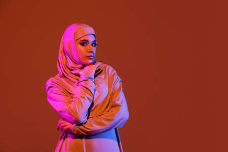Posing graceful. Beautiful arab woman in stylish hijab isolated on brown studio background in neon,...