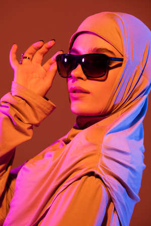 Close up. Beautiful arab woman in stylish hijab isolated on brown studio background in neon, copyspace for ad. Fashion, beauty, style concept. Female model with trendy make up, accessories.の写真素材