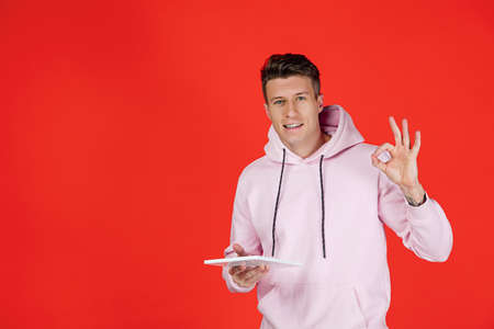 Smiling, scrolling tablet. Caucasian mans portrait isolated on red background with copyspace....