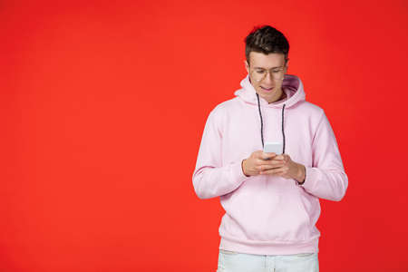 Smiling, scrolling phone. Caucasian mans portrait isolated on red background with copyspace. Handsome male model in street style. Concept of human emotions, facial expression, sales, ad, fashion.の写真素材