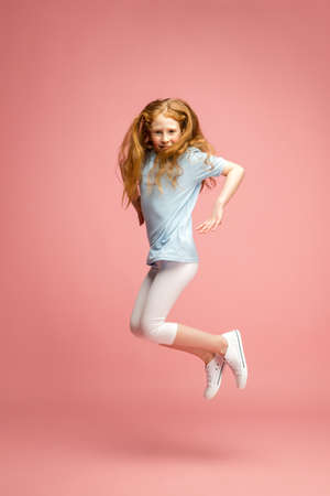 Jumping high. Happy, smiley redhair girl isolated on pink studio background with copyspace for ad. Looks happy, cheerful. Childhood, education, human emotions, facial expression concept.の写真素材