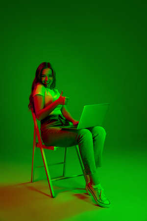 Caucasian womans portrait isolated on green studio background in multicolored neon light. Typing on laptop, thumb up. Concept of human emotions, facial expression, sales, ad, fashion. Copyspace.の写真素材