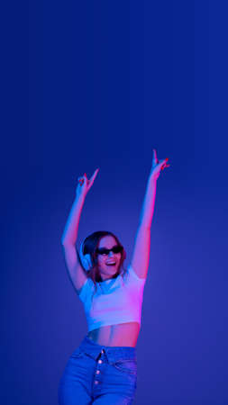 Caucasian womans portrait on blue studio background in multicolored neon light. Beautiful model with headphones. Concept of human emotions, facial expression, sales, ad, fashion. Copyspace. Flyerの写真素材