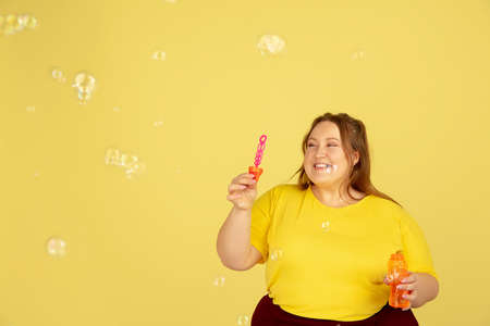 Soap bubbles. Beautiful plump caucasian plus size model isolated on yellow studio background. Concept of inclusion, human emotions, facial expression, sales, body positive. Copyspace for ad.の写真素材