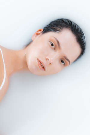 Close up female face in the milk bath with soft white glowing. Copyspace for advertising. Beauty, fashion, style, bodycare concept. Attractive caucasian model in milky colored foam.の写真素材