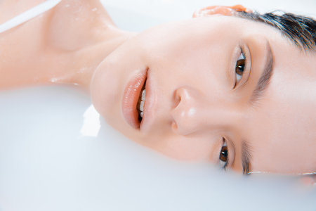 Close up female face in the milk bath with soft white glowing. Copyspace for advertising. Beauty, fashion, style, bodycare concept. Attractive caucasian model in milky colored foam.の写真素材
