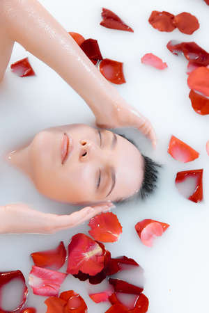 Close up female face in the milk bath with soft white glowing and rose petals. Copyspace for advertising. Beauty, fashion, style, bodycare concept. Attractive caucasian model in milky colored foam.の写真素材