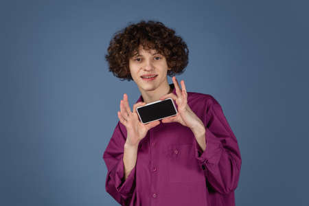 Shows blank screen. Caucasian young handsome mans portrait on blue studio background with copyspace. Male curly model with phone. Concept of human emotions, facial expression, sales, ad, fashion.の写真素材