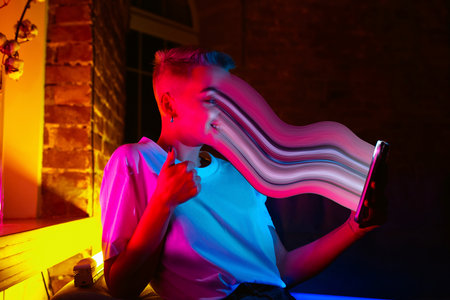 Cinematic portrait of stylish woman in neon lighted interior using a smartphone. The face is smeared, sucked, absorbed into the screen. Concept of social network dependency, phone addiction.の写真素材