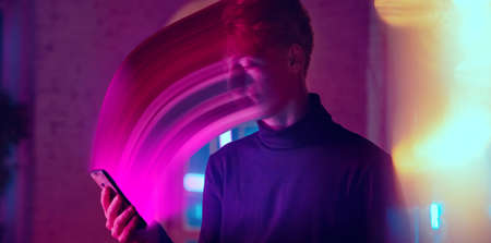 Cinematic portrait of stylish man in neon lighted interior using a smartphone. The face is smeared, sucked, absorbed into the phone. Concept of social network dependency, phone addiction.の写真素材
