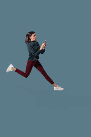 Scrolling phone on the run. High angle view of young woman on blue studio background. Human emotions and facial expressions concept. Full length portait, copyspace for ad. Fashion, retro style.の写真素材