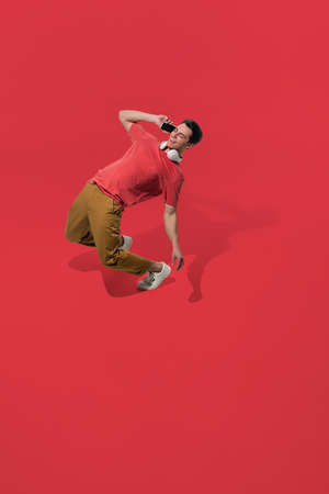Talking phone, wearing headphones. High angle view of young man on red background. Human emotions and facial expressions concept. Full length portait, copyspace for ad. Fashion, retro style.の写真素材