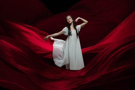 Flying. Young and graceful ballet dancer on flying red cloth background in classic action. Art, motion, action, flexibility, inspiration concept. Flexible caucasian ballerina with billowing red waves.の写真素材