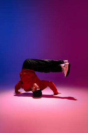 Beautiful sportive boy dancing hip-hop in stylish clothes on colorful gradient background at dance...