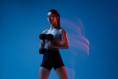 Caucasian professional female athlete training on blue studio background in neon, mixed light. Muscular, sportive woman.の写真素材
