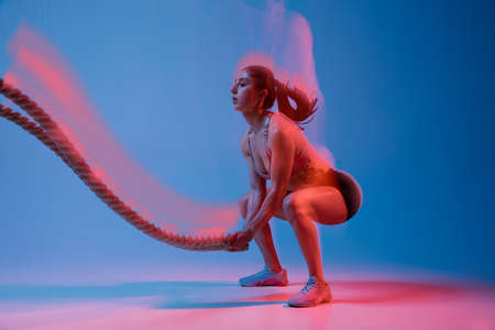 Caucasian professional female athlete training on blue studio background in neon, mixed light. Muscular, sportive woman.の写真素材