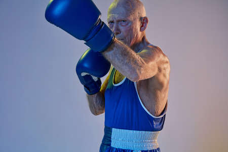 Senior man wearing sportwear boxing isolated on gradient studio background in neon light. Concept of sport, activity, movement, wellbeing. Copyspace, ad.の写真素材