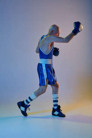 Senior man wearing sportwear boxing isolated on gradient studio background in neon light. Concept of sport, activity, movement, wellbeing. Copyspace, ad.の写真素材