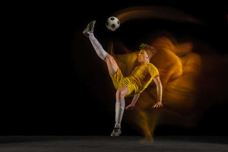 Young caucasian male football or soccer player kicking ball for the goal in mixed light on dark...