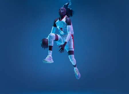 Beautiful african-american female basketball player in motion and action in neon light on blue background. Concept of healthy lifestyle, professional sport, hobby.の写真素材