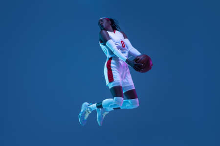 Beautiful african-american female basketball player in motion and action in neon light on blue background. Concept of healthy lifestyle, professional sport, hobby.の写真素材