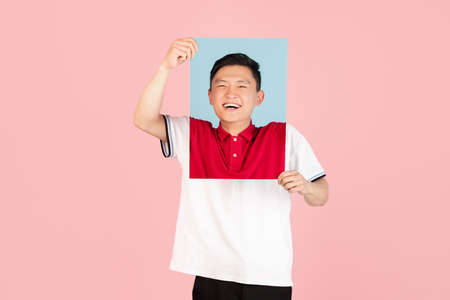 One Asian young man holding his portraits isolated on pink studio background. Concept of human...