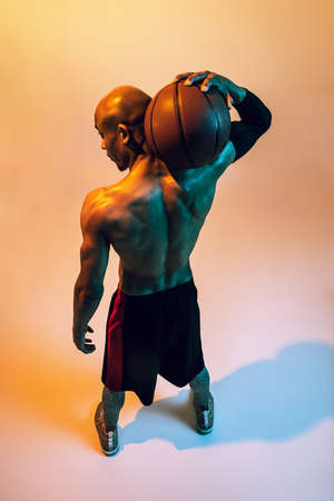 Sportive muscular african-american male basketball player posing in neon light on orange background. Back viewの写真素材