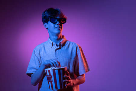 Excited in 3D eyewear and with popcorn. Caucasian mans portrait isolated on blue studio background in pink neon light. Beautiful male model.の写真素材