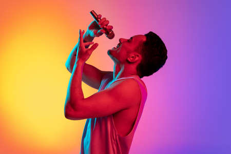 Portrait of Latina young man, singer with microphone performing isolated on gradient yellow purple background in neon light. Concept of emotions, facial expressionの写真素材