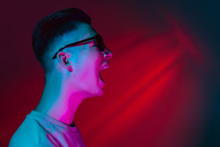 Portrait of Caucasian young man in sun glasses isolated on dark red studio background in neon pink light. Concept of human emotions, facial expression.の写真素材