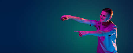 Young Caucasian man in casual clothes isolated over gradient blue green studio background in pink neon light with copyspace for ad. Concept of human emotionsの写真素材