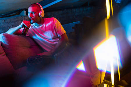 Portrait of handsome young Caucasian man sitting at home in neon orange pink lighted room. Stylish...