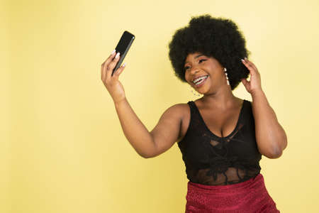 Beautiful African young woman posing with phone isolated over light yellow studio background. Concept of human emotions, facial expression, youth, sales, ad.の写真素材