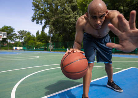 Stop, defense. Young man, muscular african male basketball player playing basketball at street public stadium, sport court or palyground outdoors. Summer sport games.の写真素材
