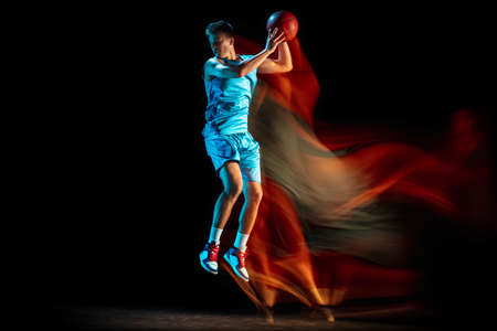 Young caucasian male basketball player playing basketball isolated over dark studio background in mixed light. Concept of healthy lifestyle, professional sport, hobby.の写真素材