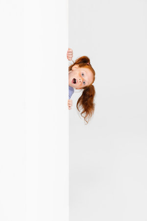 Portrait of cute freckled red-headed girl in casual outfit peeking out the wall isolated on white studio background. Happy childhood concept. Sunny childの写真素材