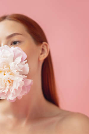 Crop portrait of beautiful redheaded woman with flower isolated over pink studio back ground. Nude color, diet, cosmetics, natural beauty and aesthetic cosmetology concept.の写真素材
