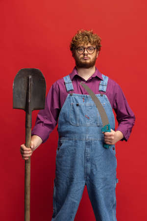 Portrait of redheaded bearded man, farmer with garden equipment isolated over red studio background. Concept of professional occupation, work.の写真素材