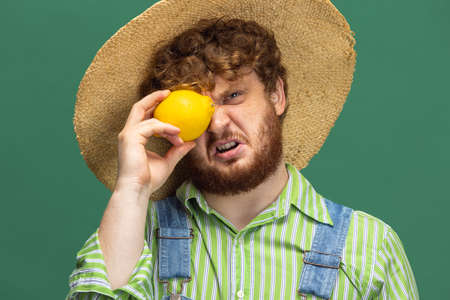 Close-up funny redheaded bearded man, farmer with yellow pepper standing isolated over green studio...