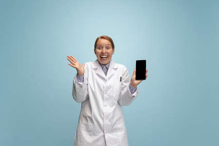Portrait of female doctor, therapeutic or medical advisor with smartphone screen isolated on blue...