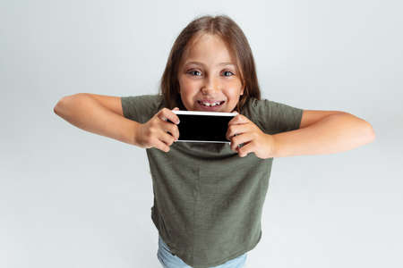 Close-up portrait of beautiful preschool smiling girl with phone isolated on white studio background. Copyspace for ad. Childhood, education, emotion conceptの写真素材